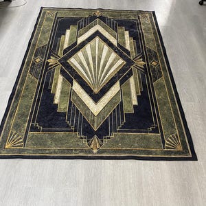 May include: A rectangular area rug featuring a dark navy blue and olive green geometric pattern. The design includes a central diamond shape with radiating fan-like lines, surrounded by layered angular borders and gold-toned accents throughout.