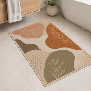 May include: A rectangular bath mat featuring an abstract pattern of organic shapes in shades of terracotta, sage green, dark brown, and cream. The design includes white line art of leaves and small dashes, with a thin border framing the entire mat.
