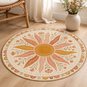 Op de afbeelding: Een rond vloerkleed met een centraal gouden zonmotief omringd door stralende roze en gele bladeren en kleine bloemtakjes. De rand bestaat uit een herhalend patroon van halfronde vormen in oranje en crèmetinten.