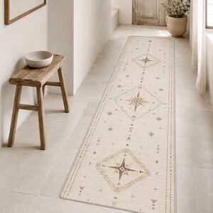 May include: A long, rectangular runner rug in a light beige color featuring a repeating pattern of geometric diamond shapes and star-like compass motifs in shades of brown and tan, with a thin border running along the edges of the fabric.