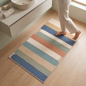 May include: A rectangular bath mat featuring a pattern of horizontal, rounded-end stripes in shades of blue, sage green, terracotta, beige, and cream. The mat has a textured surface and is positioned on a light wood floor in a bathroom setting.