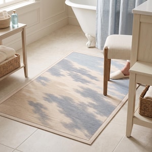 May include: A rectangular area rug featuring a soft, abstract cloud-like pattern in shades of muted blue and beige. The rug has a finished border and a flat-woven texture, positioned on a light-colored tiled floor in a bathroom setting.