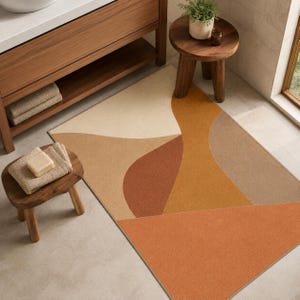 May include: A rectangular area rug featuring a geometric pattern of abstract shapes in shades of cream, beige, terracotta, and burnt orange. The rug is positioned on a light-colored floor near a wooden vanity, a small wooden stool, and a plant.