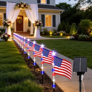 May include: A row of American flags with red and white stripes and blue star fields, illuminated by white star-shaped lights. The flags are set along a pathway, with a solar panel in the foreground. The scene is set in front of a house with a decorated entrance.