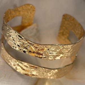 May include: A gold-colored cuff bracelet with a textured, hammered finish. The open design features two parallel bands connected by a bridge. The bracelet has a shiny, reflective surface, suggesting it is made of metal.