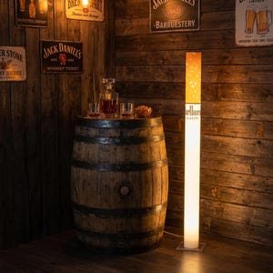 May include: A tall, cylindrical floor lamp designed to resemble a cigarette with a white base, a yellow filter section, and the text Marlboro Lights printed on the side. The lamp emits a warm glow, standing upright on a square base.
