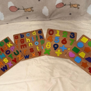 May include: Four wooden puzzle boards featuring colorful letters, numbers, and shapes. One board displays uppercase letters, another lowercase letters, a third shows numbers and math symbols, and the fourth features various geometric shapes.
