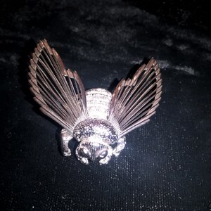 May include: A silver bee brooch with intricate details and a textured body. The wings are made of thin, delicate metal and are spread out in a fan shape.