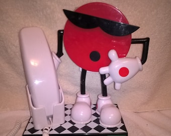 May include: A red and white 7 Up phone with a black and white checkered base. The phone features a 7 Up mascot with sunglasses and a white glove.