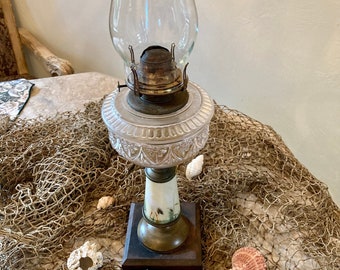 May include: A vintage oil lamp with a clear glass chimney and a frosted glass base with a decorative pattern. The base is painted with a floral design and sits on a wooden platform.
