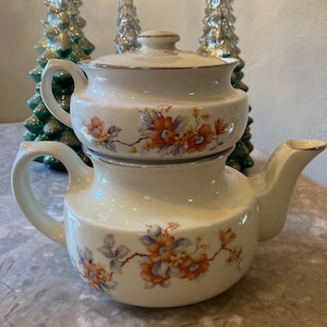 May include: A white teapot with a floral design in shades of orange, yellow, and purple. The teapot has a removable top and a handle on each side.
