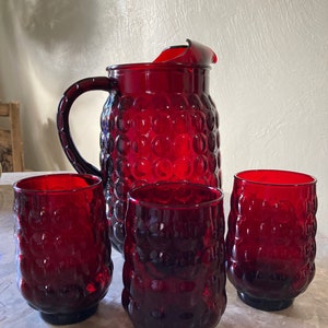 May include: A ruby red glass pitcher with a large handle and three matching drinking glasses. The pitcher and glasses have a textured, bubble-like pattern. The set is displayed on a marble-like surface.