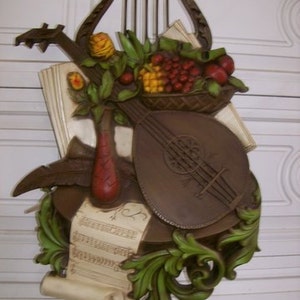 May include: A brown and gold wall sculpture featuring a lyre, a lute, a basket of fruit, and a scroll of sheet music. The sculpture is made of a textured material and has a rustic, vintage look.