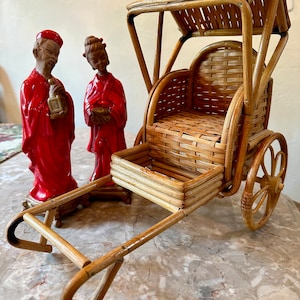 May include: A miniature bamboo rickshaw with a red and white ceramic figurine of a man and a woman in traditional clothing. The rickshaw has a woven seat and a canopy.