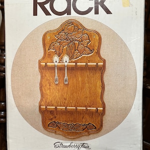 May include: A wooden spoon rack with a carved strawberry design. The rack is made by Cornwall and is called "Strawberry Fair". It is 10 inches by 1 1/4 inches by 18 inches and is made in a spring wheat finish.