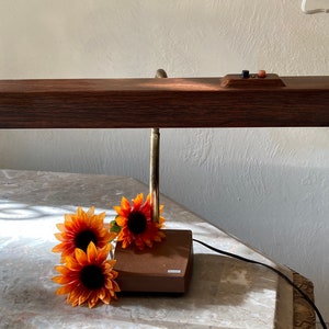 May include: A vintage wooden desk lamp with a brown wood base and a long, rectangular shade. The lamp has a brass arm and a brown cord. Three orange sunflowers are in the foreground.