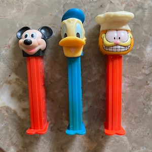 May include: Three vintage PEZ dispensers featuring cartoon characters Mickey Mouse, Donald Duck, and Garfield. Mickey has a black head and red stem. Donald Duck has a blue stem. Garfield has an orange head and stem.