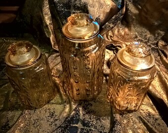 May include: Three amber-colored glass jars with ornate, textured patterns and matching lids. The jars vary in size and are arranged on a gold and black patterned fabric. The lids have a decorative, raised design.