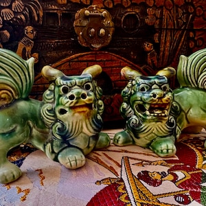 May include: Two ceramic Foo Dog statues in shades of green, with detailed manes, wings, and expressive faces. The statues are positioned on a patterned surface, with a wooden carved background.