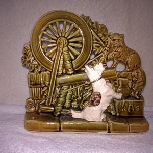 May include: A ceramic figurine of a cat and a dog, with a spinning wheel in the background. The figurine is painted in shades of green and brown, with white accents. The cat is sitting on a ledge, while the dog is standing on a platform. The spinning wheel is made of wood and has a metal spindle.