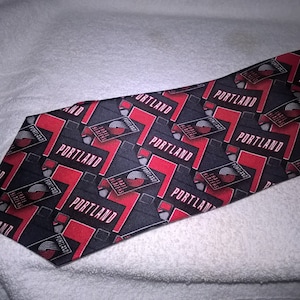 May include: A black silk tie with a repeating pattern of red and white "Portland Trail Blazers" logos. The tie has a black background with red and white text and logos.