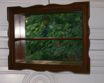 May include: A brown wooden wall shelf with a mirror and a shelf. The shelf is empty and the mirror reflects a view of green foliage.
