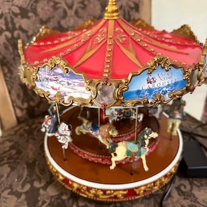 May include: A red and gold carousel with a winter scene painted on the side. The carousel has five horses and a lion, and it says "Village Square Carousel" on the side.