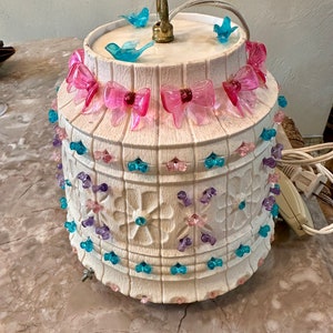May include: A white ceramic lampshade with a floral design and pink, blue, and purple plastic beads. The lampshade has a gold metal top with a hook and blue plastic birds.