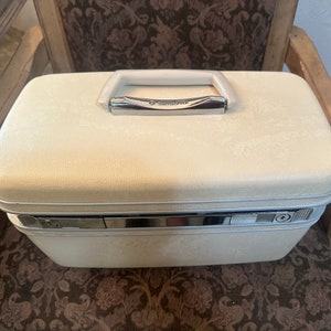 May include: A white Samsonite vintage travel case with a silver latch and handle. The case has a textured surface and is sitting on a brown patterned chair.