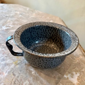 May include: A gray enamelware bowl with a speckled pattern and a single handle. The bowl is slightly chipped around the rim.