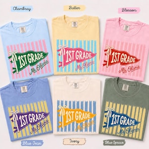 May include: Six short-sleeve t-shirts in chambray, butter, blossom, blue jean, ivory, and blue spruce colors. Each shirt features a vertical striped background and a pennant graphic with the text 1st Grade and Ms. Olivia in cursive script.