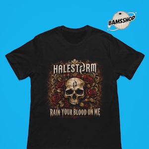 Op de afbeelding: Een zwart t-shirt met korte mouwen en een grafische print van een schedel omringd door rode rozen en goudkleurig filigraan. De tekst "HALESTORM" staat boven de schedel en "RAIN YOUR BLOOD ON ME" staat in een verweerd lettertype eronder.