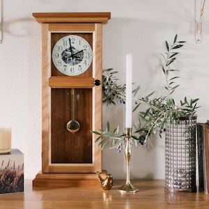 May include: A tall wooden pendulum clock with a light and dark wood frame, a white clock face featuring black numerals and a mountain illustration, a gold pendulum, a small black knob, a candle, a glass vase with olive branches, and two books.