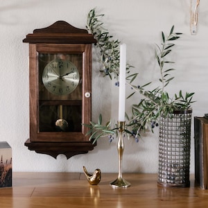 May include: A wooden wall clock with a pendulum, a candle, a gold candlestick, a decorative bird, a vase, and two books are arranged on a wooden surface. Olive branches add a natural touch to the scene.