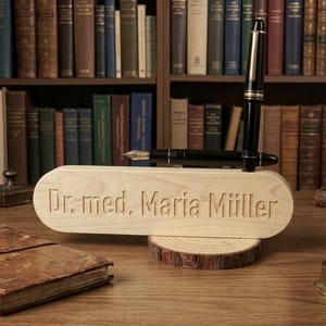 Puede incluir: Un letrero de escritorio de madera clara con el texto grabado "Dr. med. Maria Müller" que descansa sobre una base de rodaja de madera circular. Una pluma estilográfica negra con detalles dorados está colocada verticalmente detrás del letrero.
