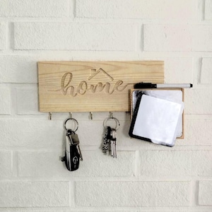 Könnte beinhalten: Schlüsselhalter aus Holz mit dem ausgeschnittenen Wort „home“, einem kleinen Dachdesign und Haken für Schlüssel. Enthält ein kleines Whiteboard mit einem schwarzen Marker. Der Schlüsselhalter ist an einer weißen Ziegelmauer befestigt.