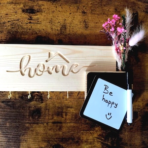 Könnte beinhalten: Holzdekoration für das Zuhause mit dem Wort "home" in das Holz geschnitzt und einer Haussilhouette. Es hat Haken für Schlüssel und einen Notizblock mit "Be happy" und einem Smiley. Ein kleiner Strauß aus rosa Blumen und weißen Federn ist ebenfalls enthalten.