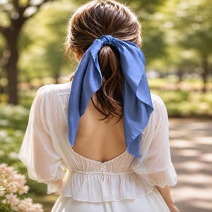 May include: A blue hair scarf tied around a ponytail, featuring a smooth, solid-colored fabric. The scarf is secured at the base of the hair, with two triangular ends draping down. The accessory is worn with a white, long-sleeved, open-back top.