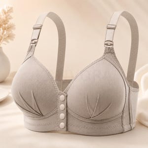 May include: A beige ribbed bra featuring a front closure with four circular buttons, adjustable shoulder straps with metal sliders, and pleated detailing at the base of each cup. The garment is constructed from a textured fabric with a solid color.