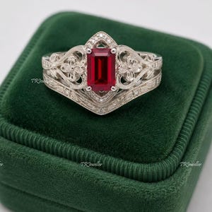 Op de afbeelding: Een zilverkleurige ring met een centrale smaragdgeslepen rode edelsteen. De band vertoont ingewikkeld filigraan metaalwerk met bloemmotieven en kleine ingebedde steentjes. De tekst TRJeweller is op meerdere plekken in de afbeelding zichtbaar.
