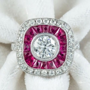 Könnte beinhalten: Ein Ring mit einem zentralen Diamanten im Brillantschliff, umgeben von einem quadratischen Heiligenschein aus tiefroten Rubinen. Ein äußerer Rand aus kleinen runden Diamanten rahmt die Rubine ein, gefasst in eine silberfarbene Ringschiene.