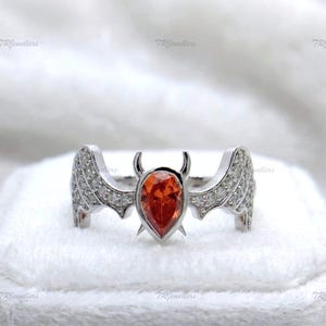 Op de afbeelding: Een zilverkleurige ring met een centrale peervormige oranje edelsteen, geflankeerd door twee vleugelvormige structuren bezet met kleine heldere steentjes. De centrale zetting heeft twee kleine hoornachtige punten aan de bovenkant.