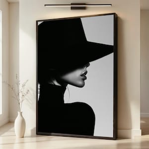 Puede incluir: Una impresión de arte mural enmarcada en blanco y negro que muestra el perfil lateral de una persona con un sombrero de ala ancha. El sombrero proyecta una sombra profunda sobre la parte superior del rostro, resaltando la nariz, los labios y la barbilla.