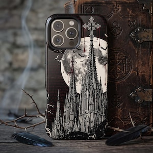 May include: A black smartphone case featuring a detailed gothic cathedral illustration under a large full moon. The design includes a tall central spire topped with a cross, flying bats, and red droplets. The case has a triple-lens camera cutout.