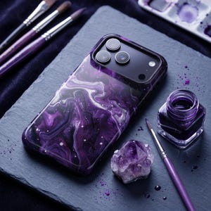 May include: A smartphone case featuring a glossy, swirling purple, black, and white marble-like pattern. The case has a cutout for a triple-lens camera system. Nearby are several paintbrushes, a small glass jar of purple ink, and an amethyst crystal.