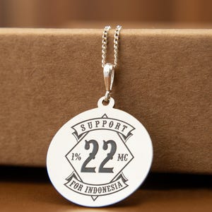 May include: A silver-toned circular pendant necklace featuring a central diamond shape with the number 22, flanked by 1% and MC. The design includes banners with the text SUPPORT and FOR INDONESIA, all suspended from a delicate silver chain.