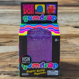Könnte beinhalten: Ein Nee Doh Gumdrop Quetschspielzeug in einer schwarzen Schachtel mit Sichtfenster. Die Verpackung zeigt bunte Wellenlinien, den Text Nee Doh gumdrop, Super Solid Squish und HSN MART LLC. Die Schachtel enthält einen Warnhinweis und 3+.