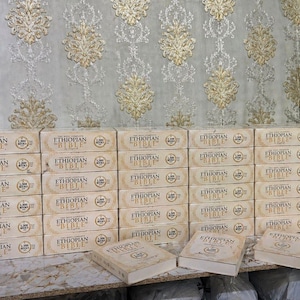 May include: Stacks of beige-colored "Ethiopian Bible" books in English. The books are in boxes with the title printed in black. The background features a patterned wallpaper with gold and beige floral designs.