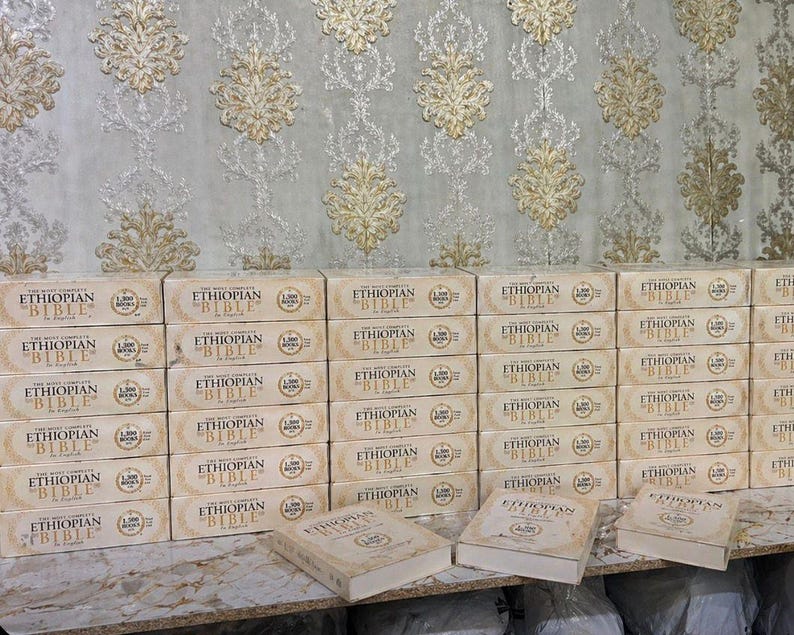 May include: Stacks of beige-colored "Ethiopian Bible" books in English. The books are in boxes with the title printed in black. The background features a patterned wallpaper with gold and beige floral designs.