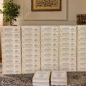 May include: A large stack of beige boxes labeled "ETHIOPIAN BIBLE" in gold lettering. The boxes are neatly arranged, with two open boxes in the foreground. The background includes a framed artwork and a small potted plant.